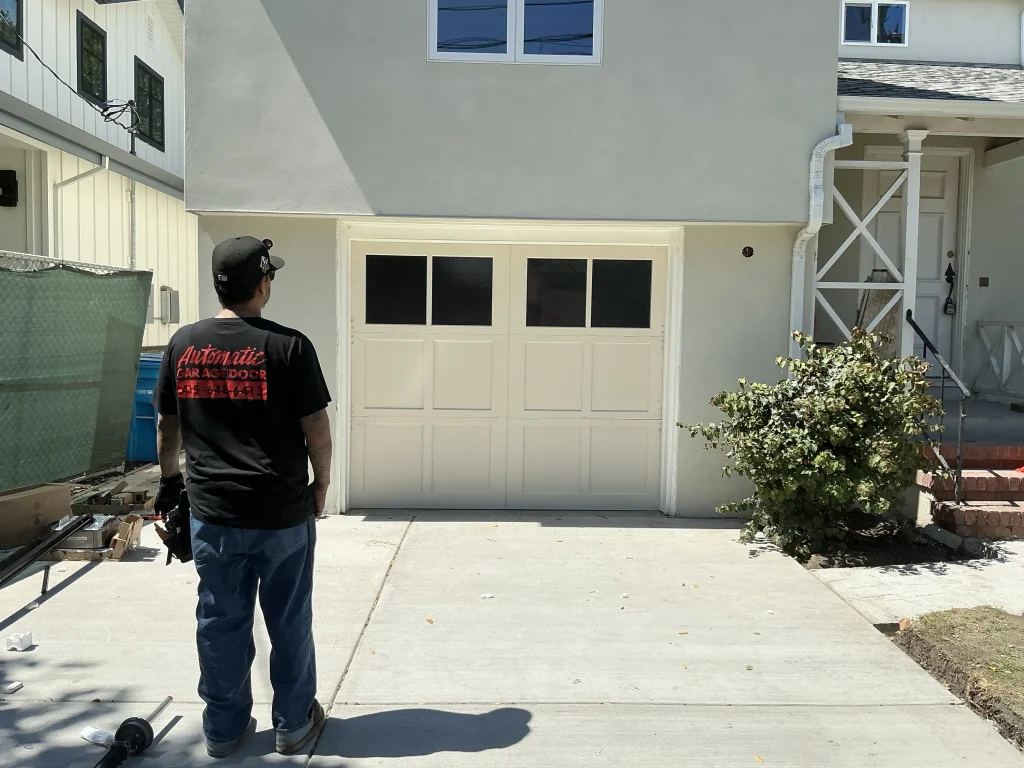 A technician standing on a driveway looking at a white sectional door with window panes during a project involving Residential Garage Doors.