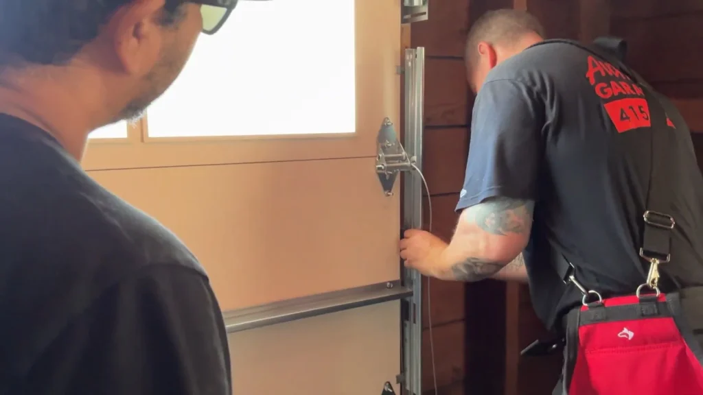 A technician repairing a door track and cable assembly while another worker observes, demonstrating residential garage door repair.