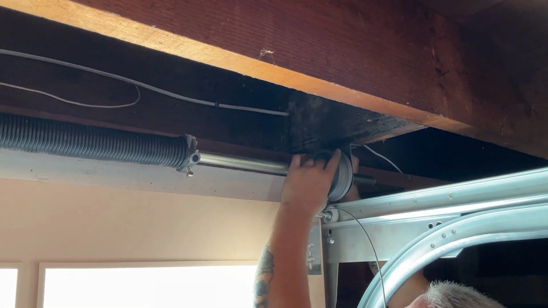 A technician reaching up to adjust a steel wire wrapping around the drum pulley during a garage door cable repair.