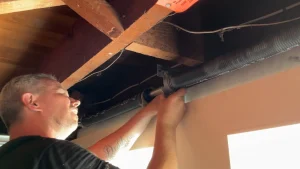 A close-up view of a technician manually working on an overhead torsion coil near wooden ceiling rafters to inspect or repair a garage door spring.
