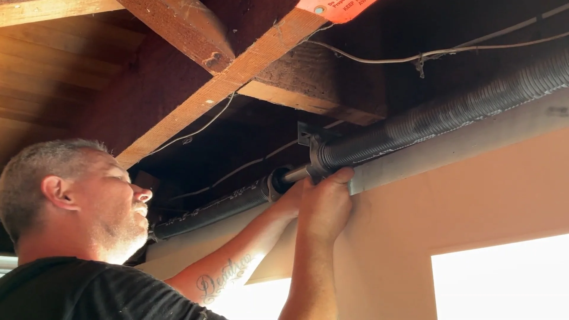 A close-up view of a technician manually inspecting an overhead torsion system near wooden rafters to address common Causes of a Garage Door Spring Break.
